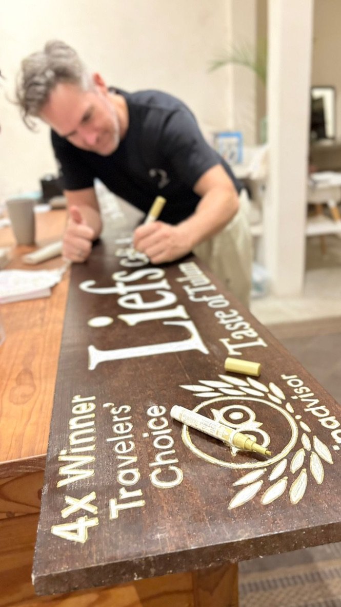 Founder Thomas hand-painting the Liefs sign on stained wood, with the Tripadvisor Travellers' Choice wreath, Rancho Verde Tulum, Tulum, Mexico