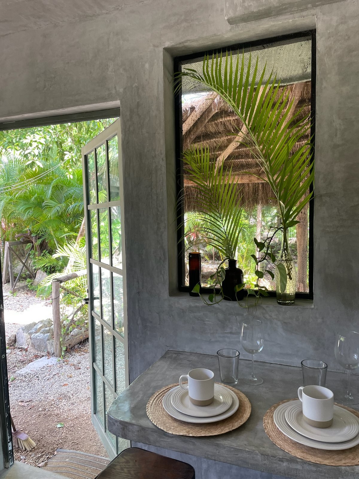 Concrete breakfast bar set for two beside an open garden door and palm-filled window, The Garden House, Airbnb at Rancho Verde Tulum