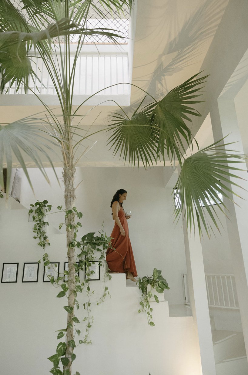 Guest in a terracotta dress walking down the white villa staircase beside an indoor palm at Rancho Verde Tulum